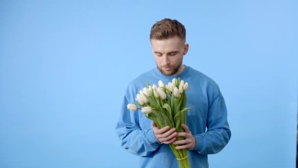 Мужчина держит букет тюльпанов, смотрит на камеру на голубом фоне. Концепция празднования - Кадры, видео