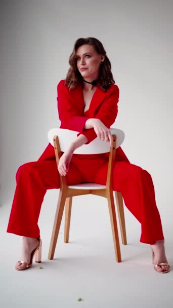 Met een introspectieve blik toont een dame haar houding zittend in een rood pak op een houten stoel, haar look aangevuld met minimalistische hakken. - Video