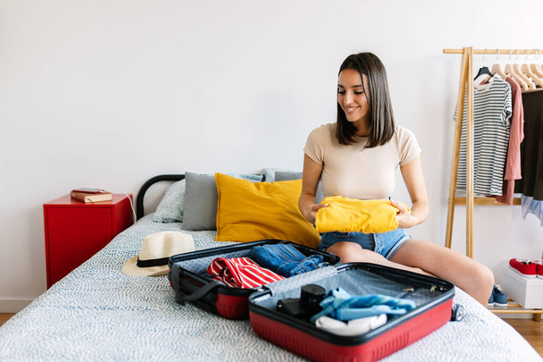 Ευτυχισμένη νεαρή όμορφη γυναίκα κάθεται στο κρεβάτι συσκευασίας βαλίτσα για ταξίδια στις καλοκαιρινές διακοπές - Φωτογραφία, εικόνα