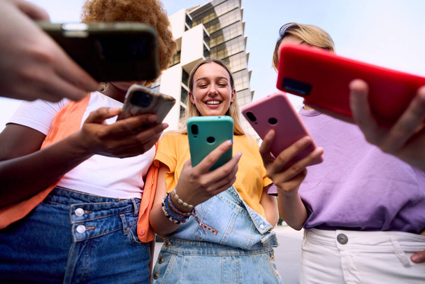 Różnorodna grupa tylko kobiet patrząc na szczęśliwy telefon komórkowy. Niski kąt widzenia przyjaciółek uzależnionych od technologii. Ludzie zgromadzeni w kółko za pomocą telefonu komórkowego. Nowe pokolenia i sieci społecznościowe komunikacji - Zdjęcie, obraz