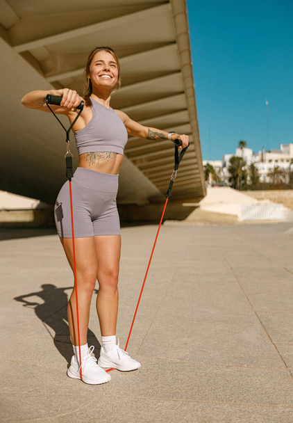 Mladá usměvavá žena dělá fitness cvičení s odporem pásmo na ranním městě pozadí - Fotografie, Obrázek