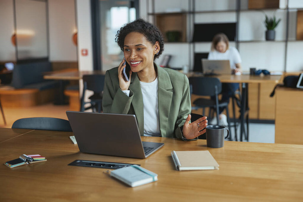 Uma mulher está sentada em uma mesa de madeira dura em um prédio, conversando em um telefone e usando um laptop. A mesa é envernizada, com uma cadeira, e piso de madeira - Foto, Imagem