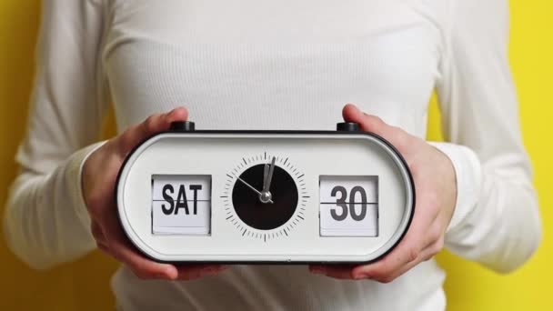 Ένα νεαρό Καυκάσιο αγνώριστο έφηβο κορίτσι που κρατάει ένα ρολόι με ώρα, ημερομηνία και μέρα: Σάββατο 30 Μαρτίου και αλλάζει σε Κυριακή 31 Μαρτίου, στέκεται μέχρι τη μέση σε ένα κίτρινο φόντο με ένα κρέμασμα - Πλάνα, βίντεο