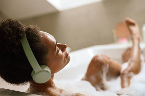 Uma mulher usando óculos está usando fones de ouvido enquanto relaxa em uma banheira. Ela está fazendo um gesto de polegar para cima com a mão como ela gosta de seu tempo de banho no quarto com acentos de madeira - Foto, Imagem