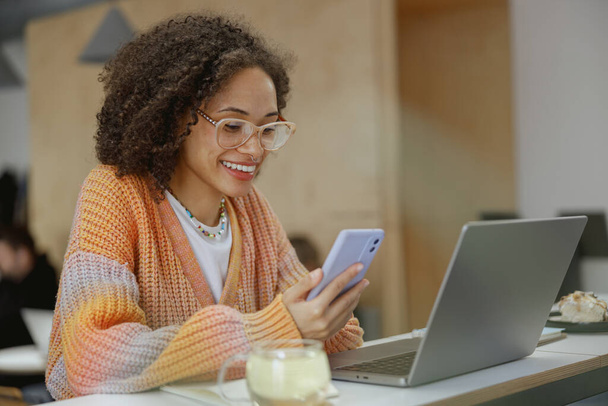 Une femme est assise à une table, souriante, utilisant un ordinateur portable et un téléphone portable. La table est encombrée de fournitures de bureau et de vaisselle - Photo, image