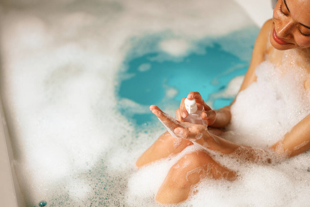 Una mujer feliz se está bañando tranquilamente en una bañera llena de agua espumosa, disfrutando de un tiempo de recreación relajante en la bañera llena de líquido - Foto, Imagen