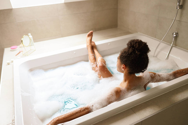 Женщина наслаждается расслабляющей ванной в ванне, наполненной пеной, в окружении успокаивающего звука воды, текущей в сантехнике из композитного материала - Фото, изображение