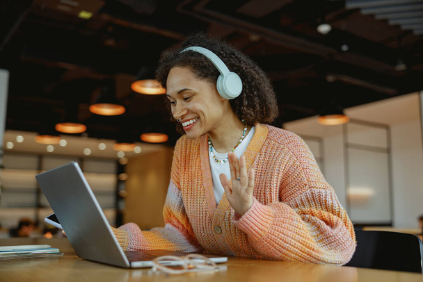 Hymyilevä nainen istuu pöydässä kannettavan tietokoneen avulla henkilökohtaisena tietokoneenaan. Hänellä on kuulokkeet ja kannettavien tietokoneiden tulostuslaite näkyy pöydällä - Valokuva, kuva