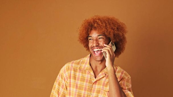 Laughing stylish curly guy speaks emotionally on mobile phone, isolated on brown background in the studio - 写真・画像