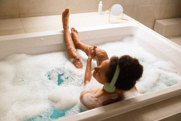 Μια γυναίκα απολαμβάνει ένα χαλαρωτικό μπάνιο σε μια μπανιέρα γεμάτη με αφρώδες νερό σε ένα μπάνιο με ξύλινα πατώματα και υδραυλικά εξαρτήματα - Φωτογραφία, εικόνα