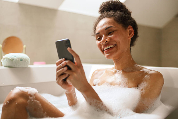 フォーマルウェアの女性は,彼女の携帯電話を見ながら喜んで風呂に入っています. 彼女は親指で笑顔とジェスチャー,おそらく楽しみのための結婚式のイベントの詳細やブライダルアクセサリーをチェック - 写真・画像