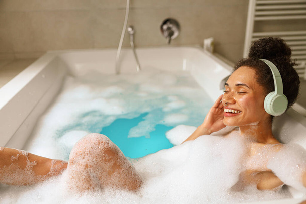 Жінка щасливо купається в ванні, наповненій водою, носить навушники, насолоджуючись неквапливим і веселим моментом у ванній кімнаті, з посмішкою на обличчі - Фото, зображення