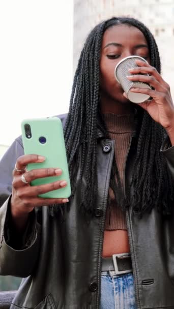 Vertical. Joven afroamericana con trenzas viendo videos en las redes sociales usando un smartphone bebiendo un café expreso parado al aire libre. Concepto de comunicación. En cámara lenta. Alta calidad 4k - Metraje, vídeo