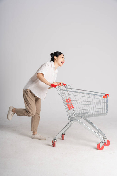 Улыбающаяся женщина с радостью толкает тележку супермаркета, изолированную на белом фоне - Фото, изображение
