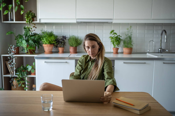 Pensive writer woman working new book nearby diary with notes ideas sitting at cozy home with indoor plants. Interested relaxed female freelancer sitting with laptop table surrounded by houseplants. - Photo, Image