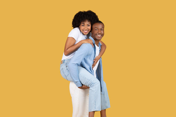 Verspieltes afroamerikanisches Paar, huckepack unterwegs, die Frau in weißem Oberteil und hellblauen Jeans, der Mann, der sie lächelnd vor leuchtend gelbem Hintergrund trägt - Foto, Bild