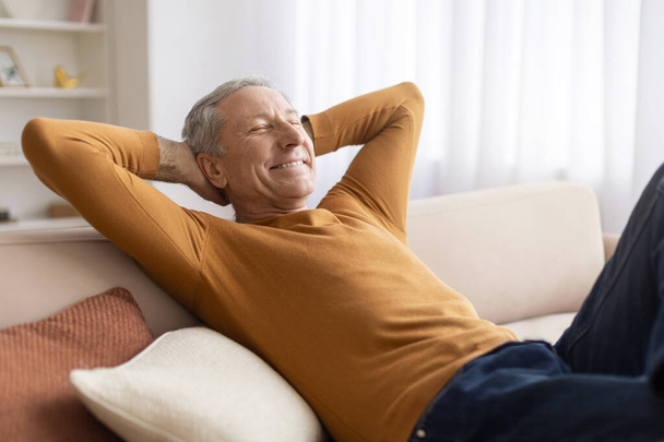 Hombre mayor relajado reclinado en el sofá en casa, sosteniendo los brazos detrás de la cabeza y sonriendo con los ojos cerrados, pensando en venir de vacaciones o fin de semana, espacio para copiar - Foto, Imagen