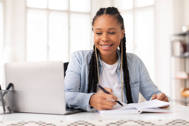 Sonriente joven estudiante negra con trenzas escribiendo notas, estudiando con computadora portátil en casa, teniendo una lección en línea mientras está sentada en una habitación luminosa - Foto, imagen