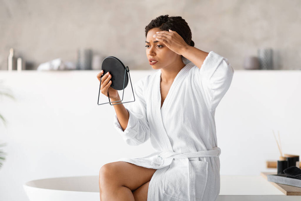 Jonge zwarte vrouw in witte badjas met spiegel, kritisch onderzoek van haar huid op het voorhoofd, bezorgd Afrikaans-Amerikaanse vrouw opgemerkt leeftijd verandert of puistjes op het gezicht, zitten op de rand van de moderne badkuip - Foto, afbeelding