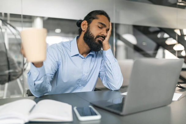 Śpiący brodaty Indianin noszący koszulę pracownik biurowy siedzący przy biurku przed laptopem z zamkniętymi oczami w biurze, trzymający kubek z kawą, wyczerpany, przestrzeń na ksero - Zdjęcie, obraz