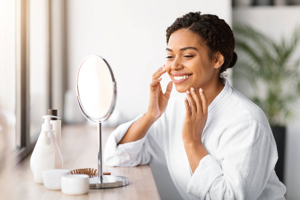 Boldog fiatal fekete nő fehér fürdőköpenyben alkalmazása arc hidratáló, miközben nézi tükröződés smink tükör, mosolygós afro-amerikai hölgy ül asztalnál körülvéve kozmetikai termékek - Fotó, kép