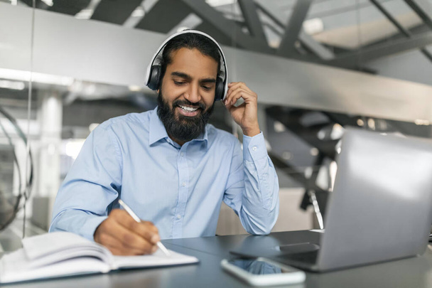 Усміхнений бородатий тисячолітній працівник індійського хлопця має онлайн-навчання, сидячи за столом в сучасному офісі, використовуючи гарнітуру і ноутбук, роблячи нотатки. Концепція професійної освіти - Фото, зображення