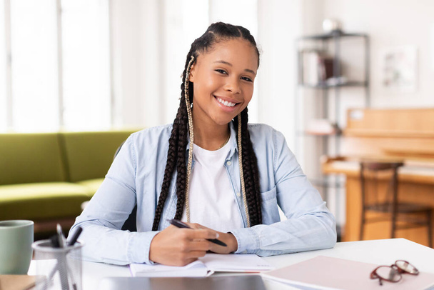 Iloinen afrikkalainen amerikkalainen naispuolinen opiskelija, jolla on punokset hymyilee, kun hän kirjoittaa muistikirjaan, istuu pöydässä kannettavan tietokoneen kanssa, huokuu rentoa opiskelutunnelmaa kotona. - Valokuva, kuva