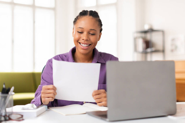 Весела чорна леді-підліток у фіолетовій сорочці радісно читає документ, сидить за її робочим столом з відкритим ноутбуком і навчальними матеріалами навколо - Фото, зображення