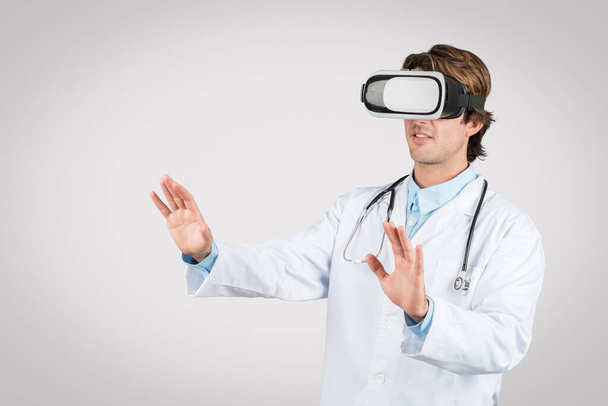 Medico maschio in camice bianco con auricolare in realtà virtuale, che fa gesti con le mani come se interagisse con la simulazione medica futuristica, sfondo grigio - Foto, immagini