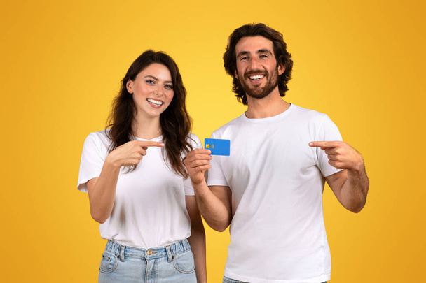 Glimlachend jong stel in casual witte t-shirts, de vrouw die naar een blauwe creditcard wijst en de man die ernaar toe gebaart, met een felgele achtergrond die symbool staat voor financiële vrijheid en geluk - Foto, afbeelding