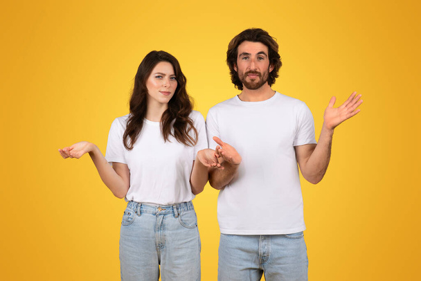 Kafası karışmış Avrupalı genç çift ellerini kaldırmış, belirsizlik ve sorgulama hareketleri sergiliyor, beyaz bluz ve kot giymiş, parlak sarı bir arka plan, stüdyo - Fotoğraf, Görsel