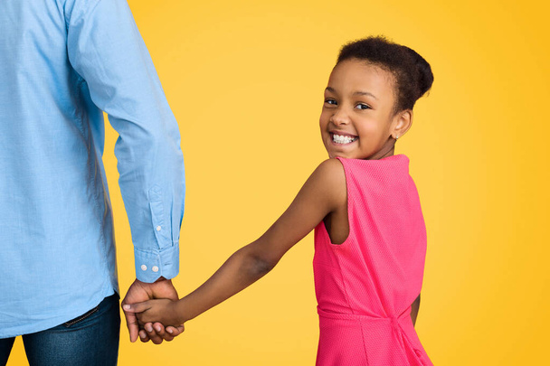 Afro-Amerikaanse meisje holding vaders hand en camera kijken op gele achtergrond - Foto, afbeelding