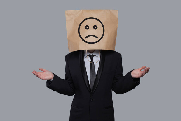 Stehender Geschäftsmann Papiertütenkopf mit traurigem Emoticon auf grauem Hintergrund - Foto, Bild