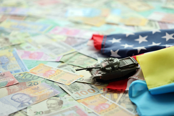 Toy tank on US flag and ukrainian flag on many banknotes of different currency. Background of war funding and military support between United States and Ukraine - Photo, Image