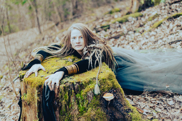 Female shaman in pristine forest nature laying on  tree stump, gold jewelry, shaggy hair - Photo, Image
