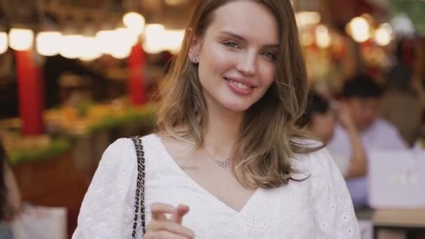Een vrouw in formele kleding, met een wit shirt, glimlacht vrolijk en zwaait naar de camera. Druk aziatische voedsel markt achter haar rug. - Video