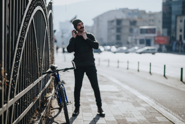 Elegante giovane adulto che utilizza uno smartphone accanto alla sua bicicletta su un marciapiede soleggiato della città, mostrando lo stile di vita urbano. - Foto, immagini