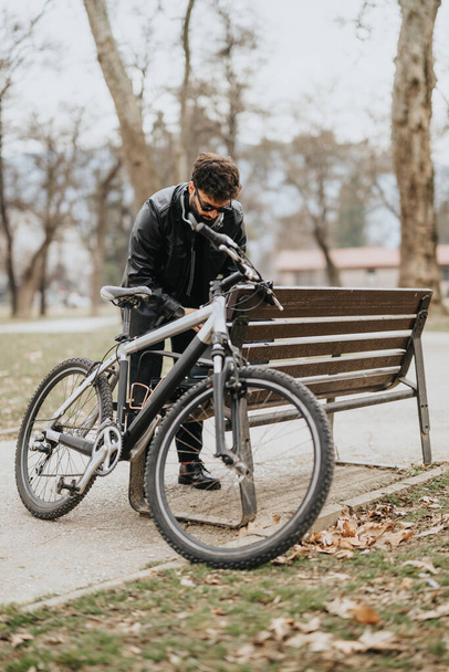 Deri ceketli bir erkek girişimci, iş ve yaşam tarzının bir karışımını sergileyerek bisikletini park bankının yanında ayarlamak için ara veriyor.. - Fotoğraf, Görsel