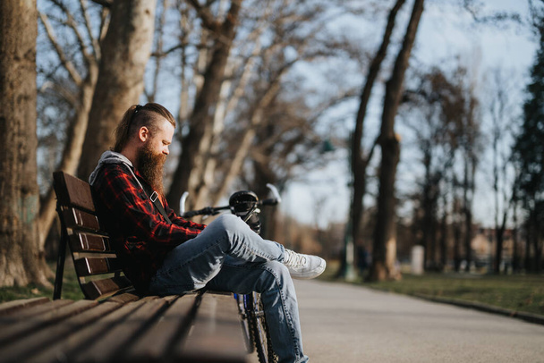 Hipster επιχειρηματίας με γενειάδα χρησιμοποιώντας ένα φορητό υπολογιστή, ενώ κάθεται σε ένα παγκάκι σε ένα αστικό πάρκο με το ποδήλατό του δίπλα του. - Φωτογραφία, εικόνα