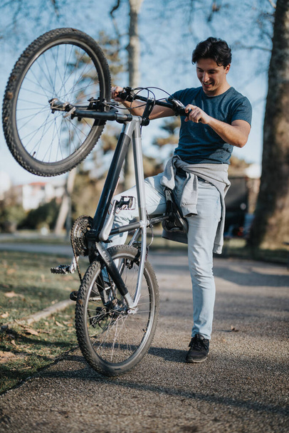 Um homem alegre gosta de recreação ao ar livre, levantando sua bicicleta de montanha em um parque com árvores no fundo. - Foto, Imagem