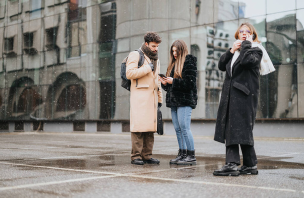 Νέοι συνάδελφοι επιχειρήσεων επικεντρώθηκε σε μια έξυπνη οθόνη του τηλεφώνου, ενώ στέκεται έξω σε μια χιονισμένη αστική μέρα, παραμένοντας συνδεδεμένοι και παραγωγικοί. - Φωτογραφία, εικόνα