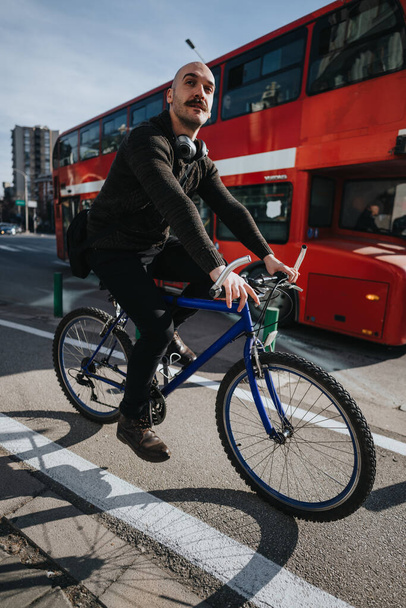 Miejski rowerzysta ze słuchawkami zatrzymujący się na skrzyżowaniu miasta z kultowym czerwonym dwupiętrowym autobusem za nim. - Zdjęcie, obraz