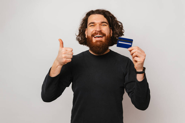 Волнующий бородатый мужчина держит синюю кредитку, показывая большой палец на белом фоне. - Фото, изображение