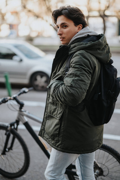 Случайный молодой человек с рюкзаком стоит рядом с велосипедом в солнечный осенний день в городе, глядя в сторону с созерцательным выражением. - Фото, изображение