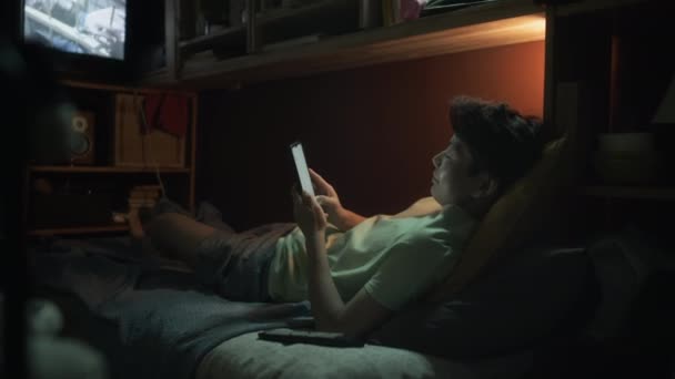 Полный снимок китайского студента в футболке и шортах, лежащих на двухъярусной кровати в тесной квартире обувной коробки и печатающих на смартфоне, фильм ужасов по телевизору, во время отдыха вечером после работы - Кадры, видео