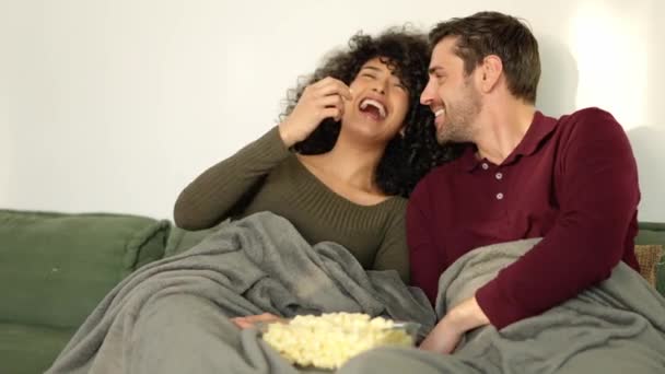Slow motion video of a multi-ethnic couple flirting and laughing eating popcorn watching movie at home - Footage, Video