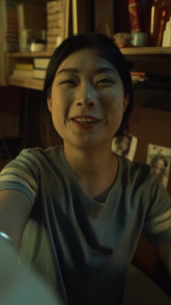 Selfie verticale medio primo piano di una ragazza asiatica sorridente che fa videochiamate con amici o familiari su smartphone, saluta, condivide notizie, ride, mentre è seduta in un piccolo dormitorio in serata - Filmati, video