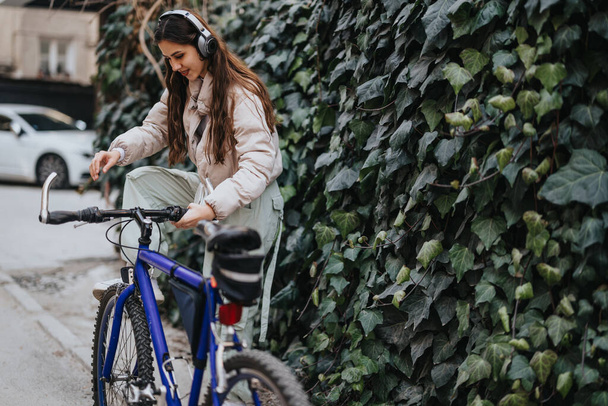 Casual młoda kobieta ze słuchawkami za pomocą smartfona na zewnątrz obok jej rower na bujnym tle bluszcz. - Zdjęcie, obraz