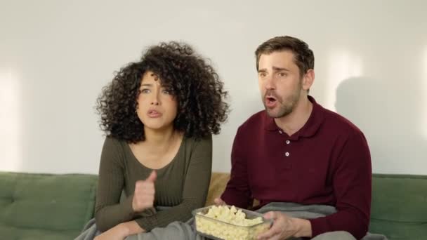 Slow motion video of a multi-ethnic couple celebrating that their football team win a match while eating popcorn lying on the sofa at home - Footage, Video