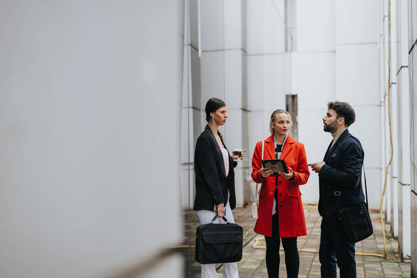 Een kleine groep professionals bespreekt werkonderwerpen terwijl ze digitale tablets en koffiekopjes buiten een modern gebouw vasthouden. - Foto, afbeelding